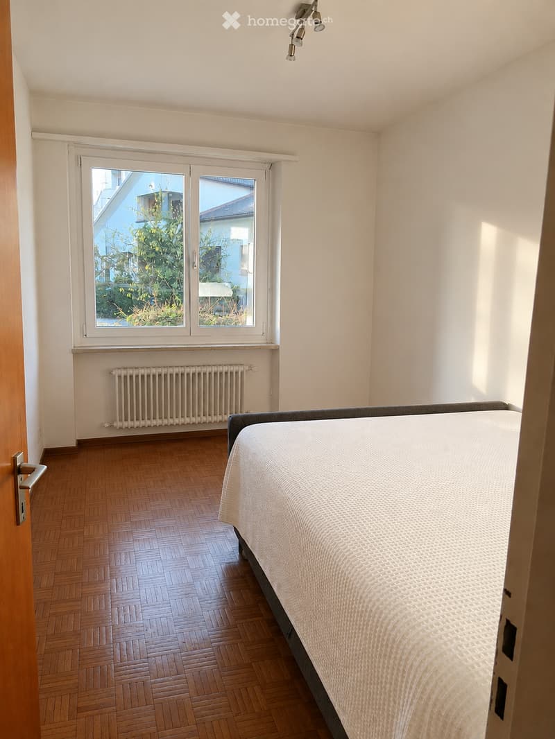 Bright bedroom with wooden herringbone flooring, a single bed with white bedding, a radiator below a large window, and a door on the left side.