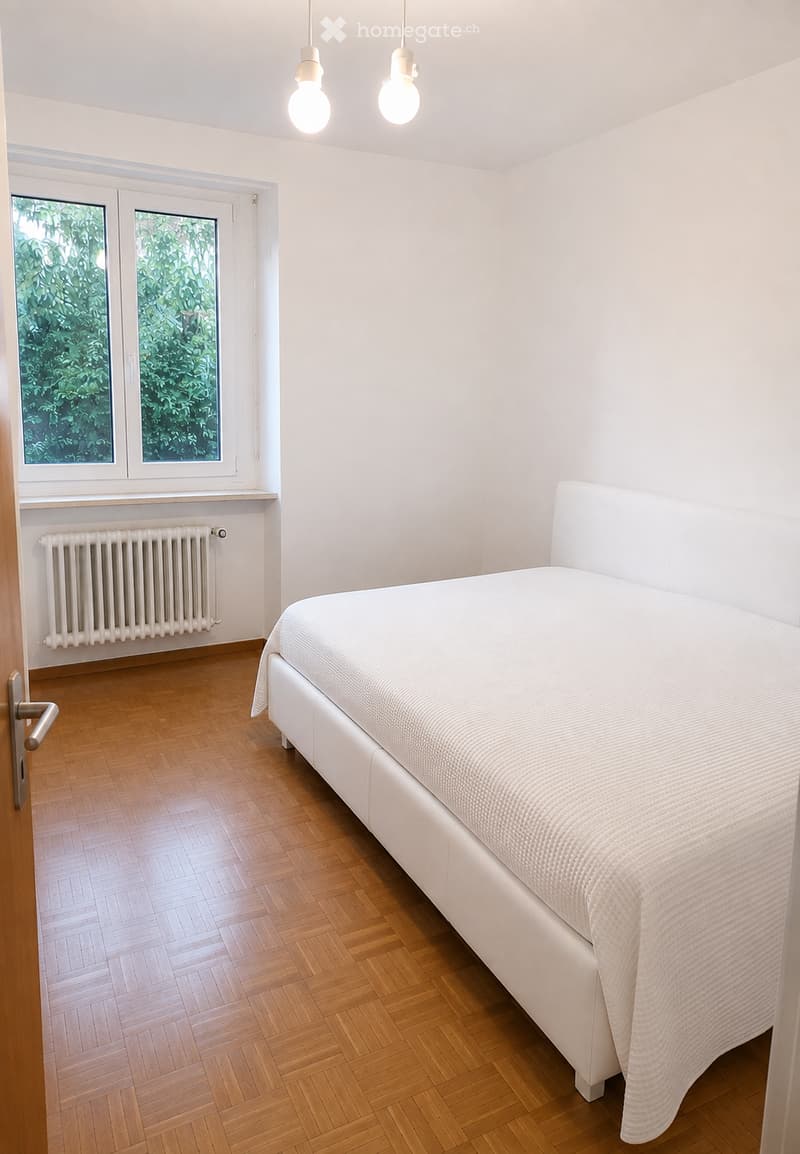 Minimalist bedroom with a white bed, wooden herringbone floor, radiator, and large windows with greenery view.