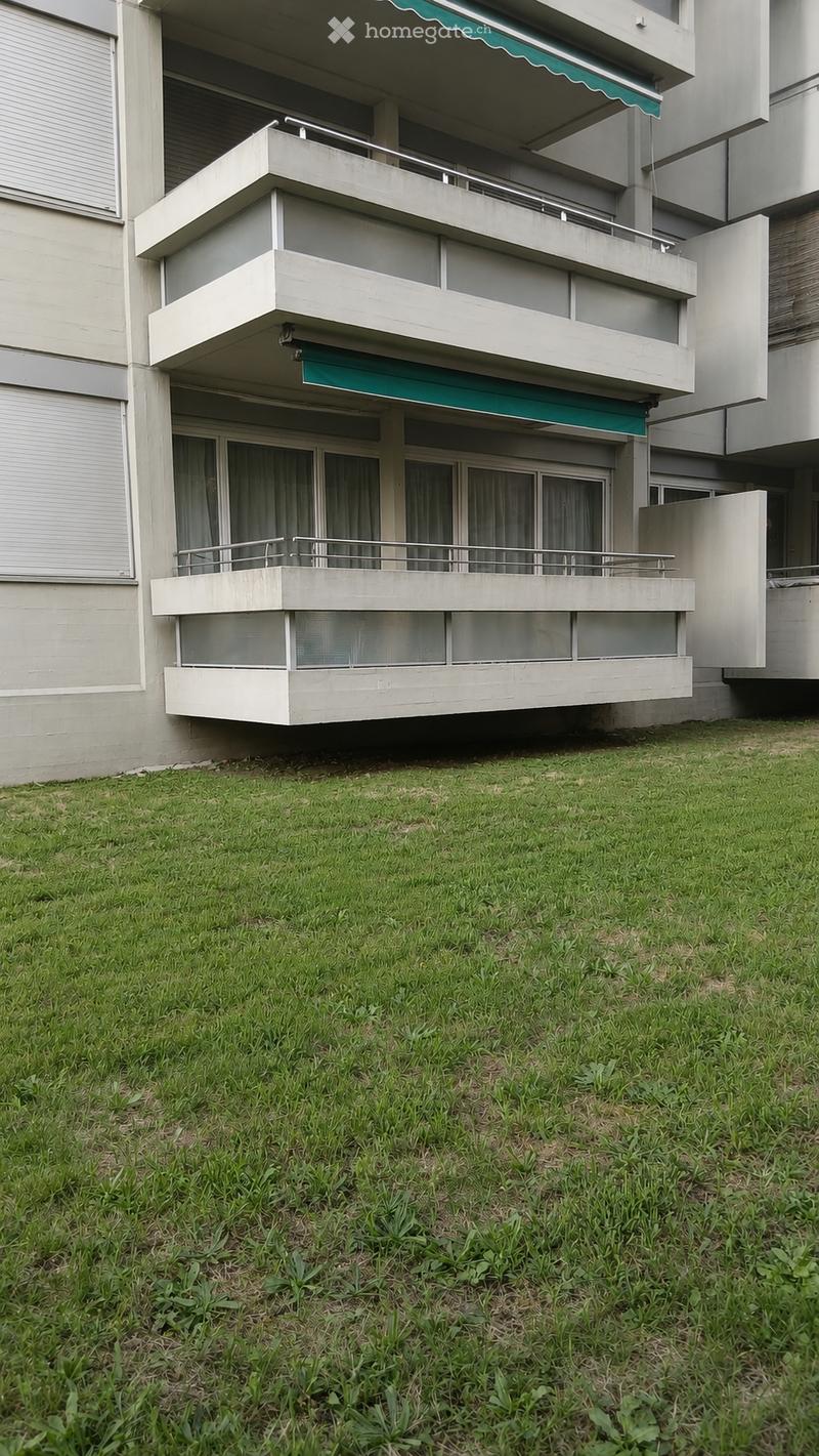 Modern multi-story building with balconies, green awnings, and large windows. The lower level has a grassy area with some plants.