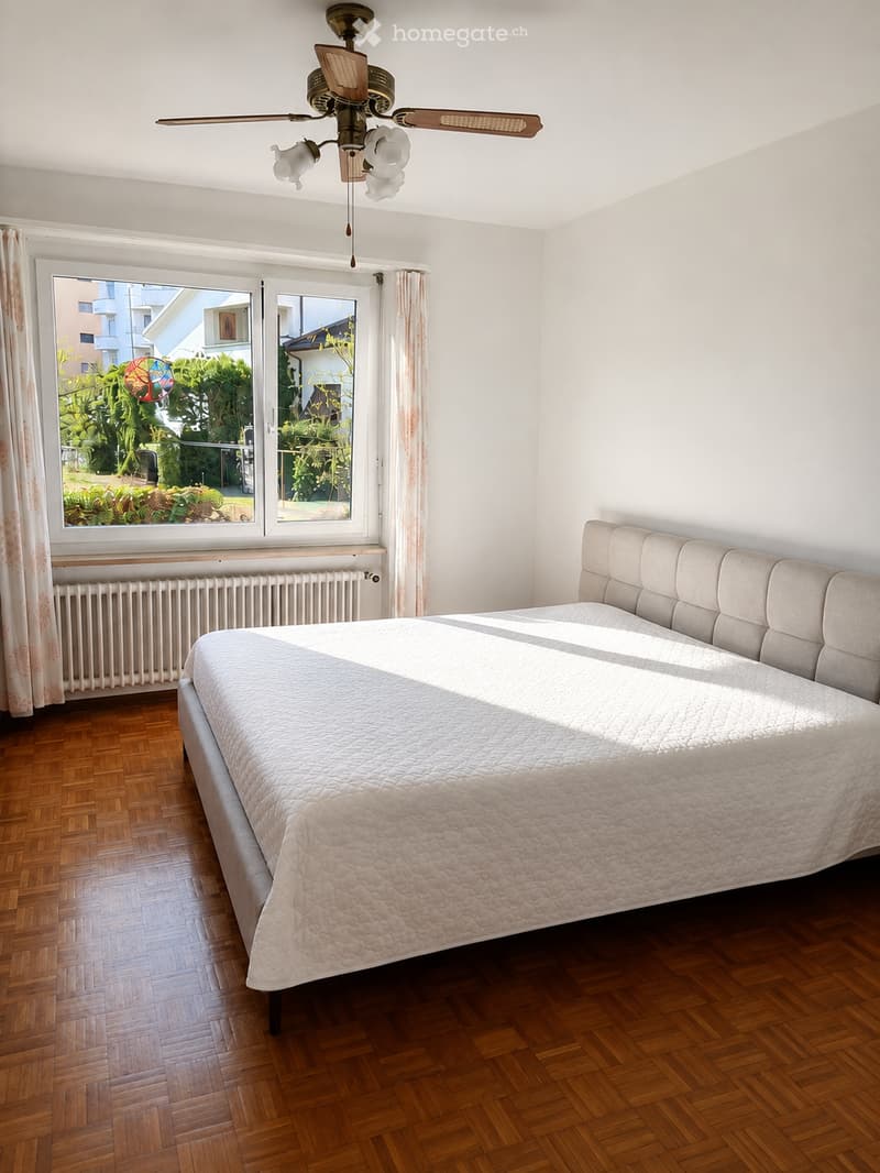 Bright bedroom with wooden flooring, large window with curtains, and a neatly made bed with a light-colored duvet and headboard.
