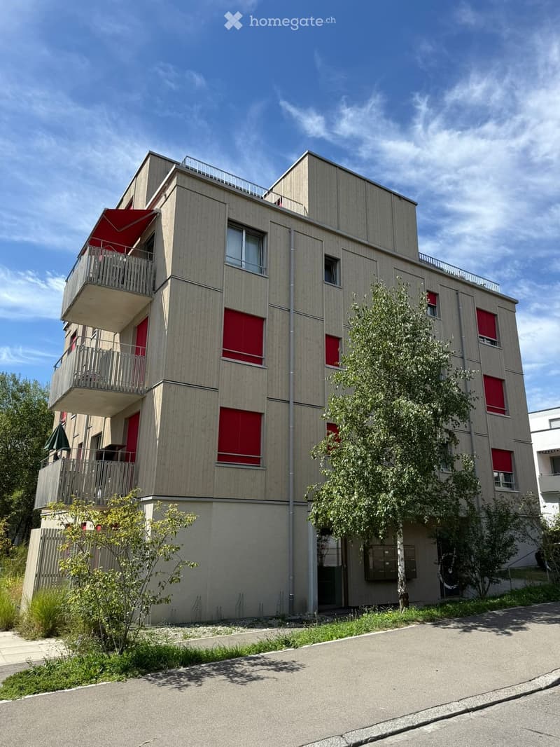 Modern three-story building with light wood siding, red shutters, balconies, and a small garden area.