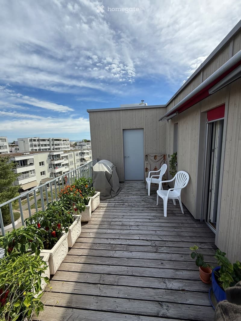 Wooden balcony with two white plastic chairs, several flower pots with plants, and a covered barbecue.