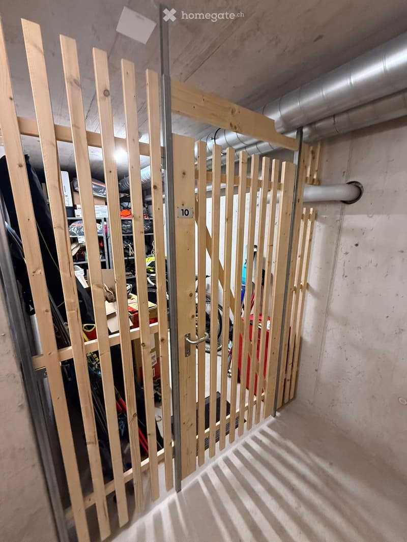 A narrow hallway with a wooden gate and a partially open door leading to a storage area. The walls are concrete, and the floor is light-colored.