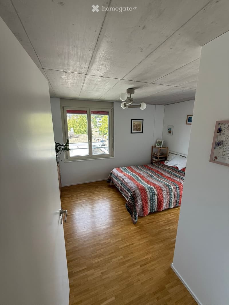 Bright bedroom with wooden flooring, a single bed with a colorful striped duvet, a nightstand with a lamp, and several picture frames on the walls. Large windows offer a view of greenery outside.