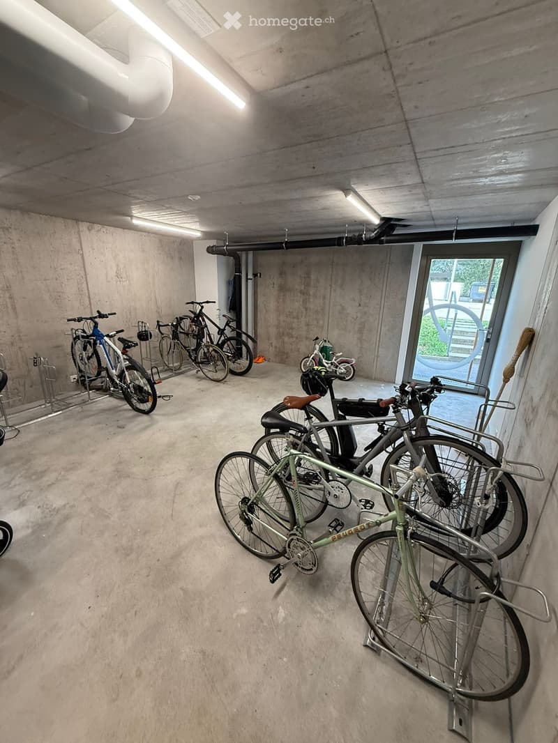 Underground parking area with several bicycles stored on racks, concrete walls and floor, and a large window showing greenery outside.