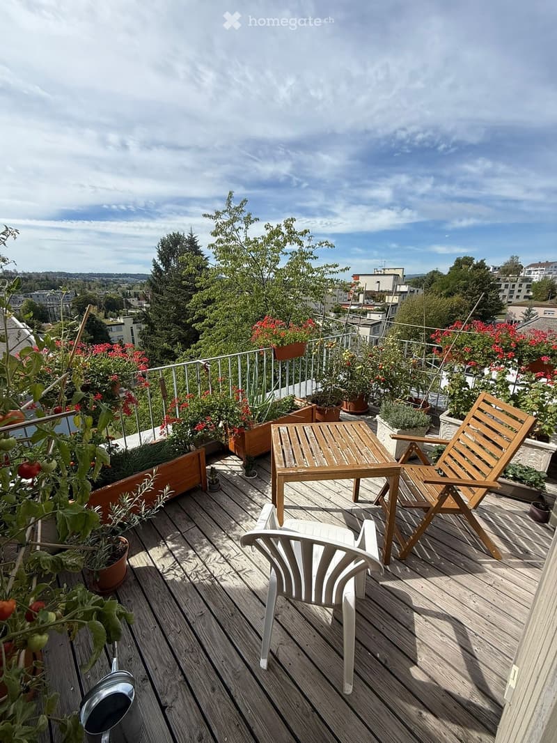 Wooden deck with a white plastic chair, wooden table, wooden folding chair, and several potted plants, overlooking a cityscape with trees and buildings.