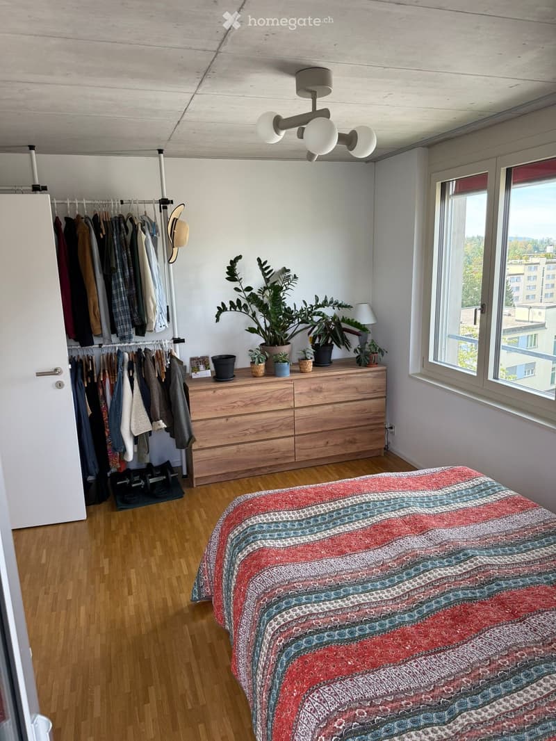 A modern bedroom with a wooden bed, colorful bedspread, dresser with potted plants, clothing rack, and a large window offering a city view.