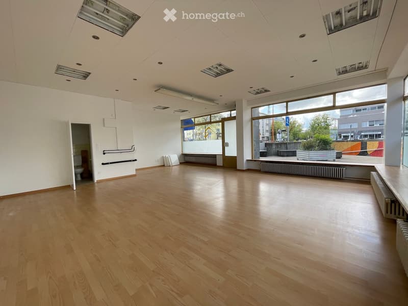 Empty room with wooden flooring, white walls, large windows, and a visible bathroom entrance.