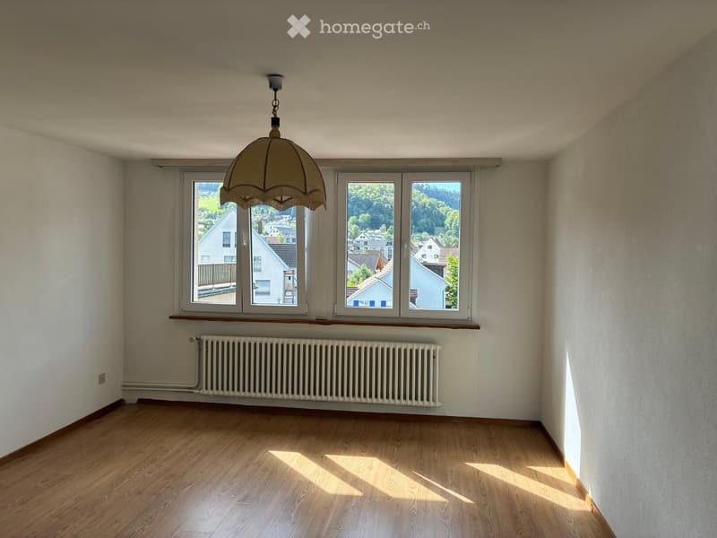 Empty room with wooden flooring, large windows offering a view of the neighborhood, a radiator, and a hanging light fixture.