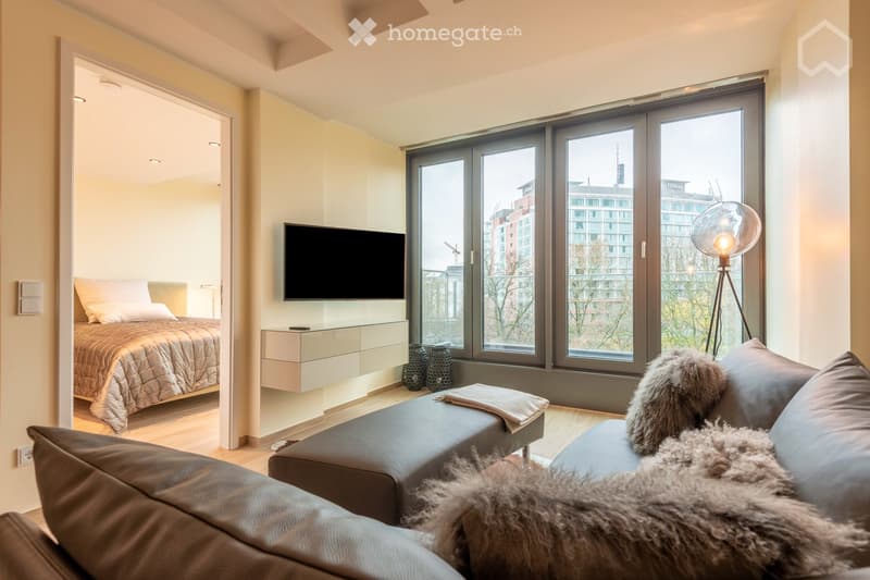 Modern living space with a sofa, bed, television, nightstand, floor lamp, and decorative vases. Large windows offer a city view.