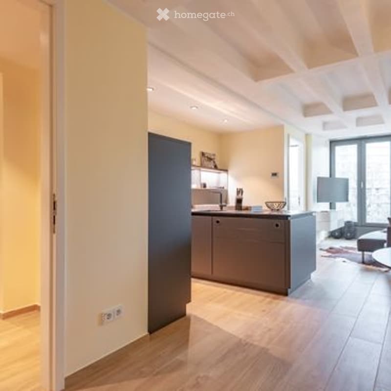 Modern open-plan living and kitchen area with light wood flooring, grey cabinetry, a central island, and a small seating area with a chair and screen.