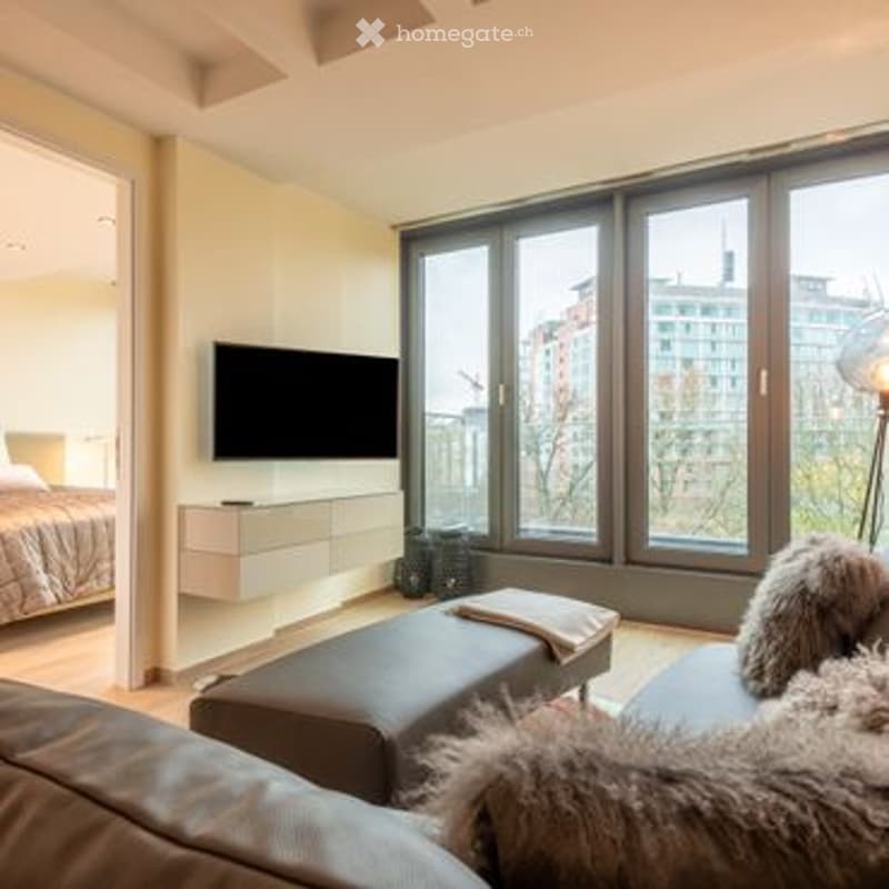 Modern living room with large windows offering a city view, a grey sofa with an ottoman, a coffee table, a flat-screen TV mounted on the wall, and a bed visible in the background.