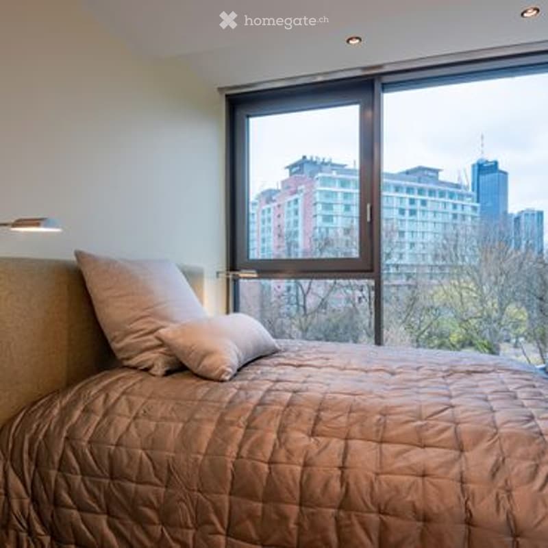 Modern bedroom with a large window offering a city view, featuring a neatly made bed with pillows and a textured bedspread.