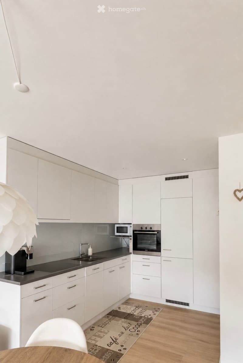 Modern kitchen with white cabinets, stainless steel appliances, a coffee maker, and a small dining area with a wooden table and white chair.