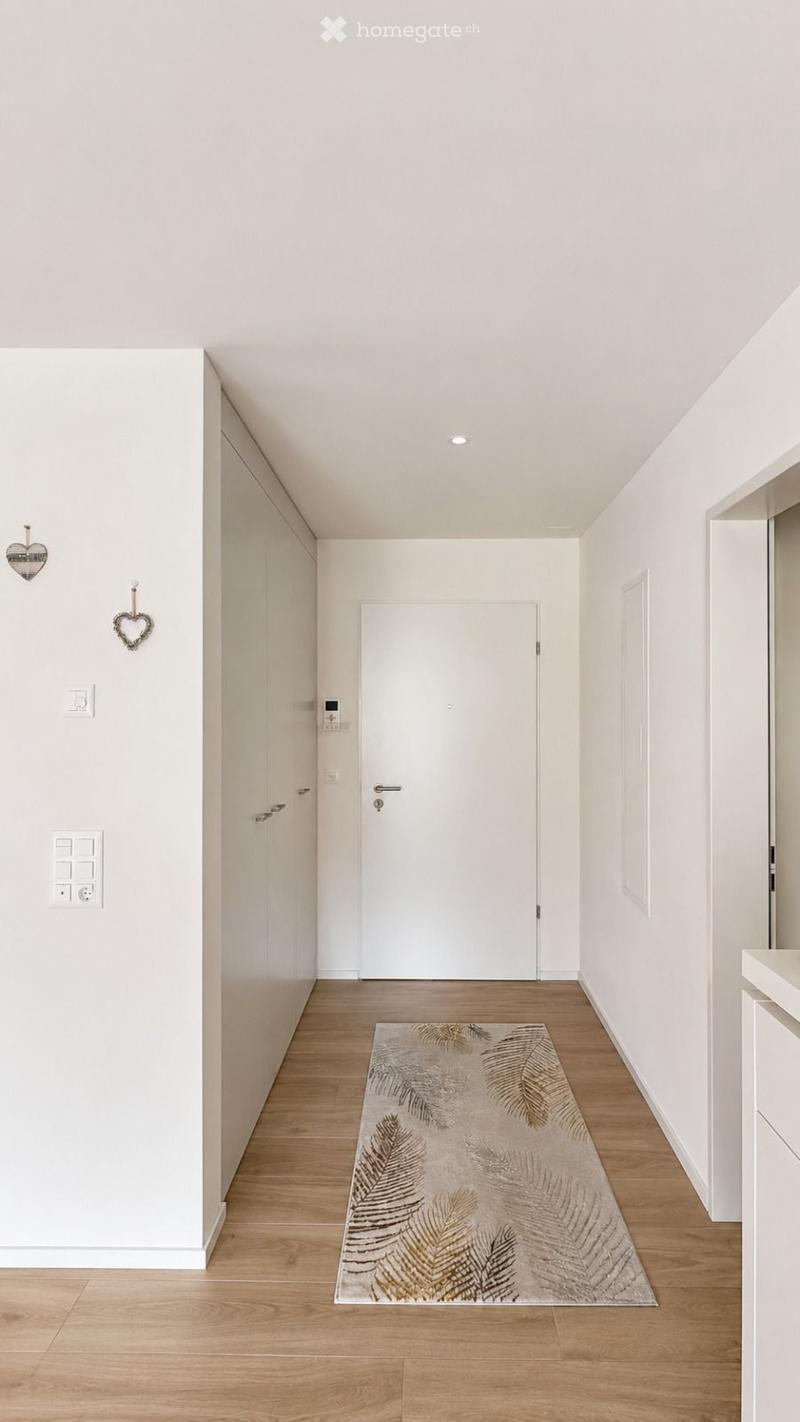 A modern, minimalist hallway with light wooden flooring, white walls, and a single door at the end. A decorative rug with leaf patterns lies on the floor, and there are built-in white cabinets on one side.