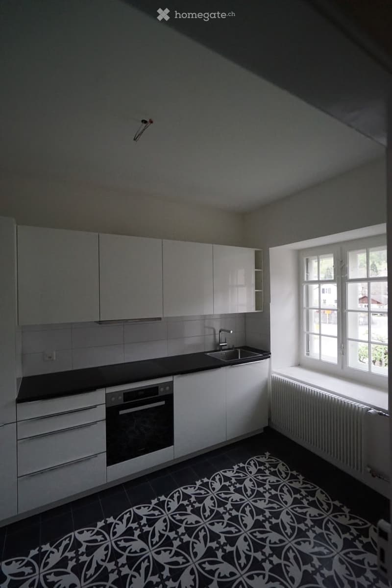 Modern kitchen with white cabinets, black countertops, and a decorative black and white tile floor. Features a sink, oven, and ample natural light from a large window.