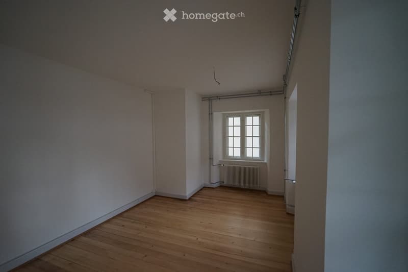 Empty room with wooden flooring, white walls, and a single window with white frames. A radiator is visible beneath the window.