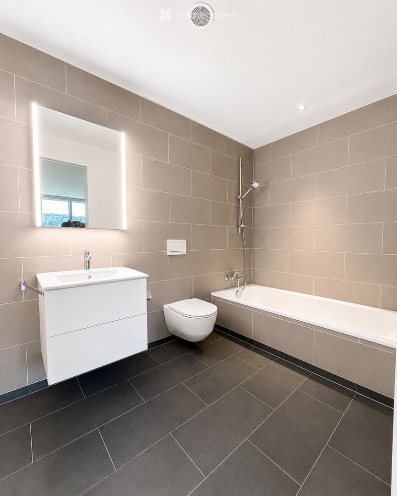 Modern bathroom with beige tiled walls, dark grey tiled floor, white sink, toilet, bathtub, and illuminated mirror above the sink.