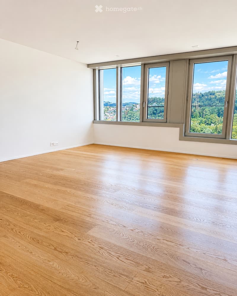 Empty room with light wooden flooring and large windows offering a scenic view of greenery and buildings.