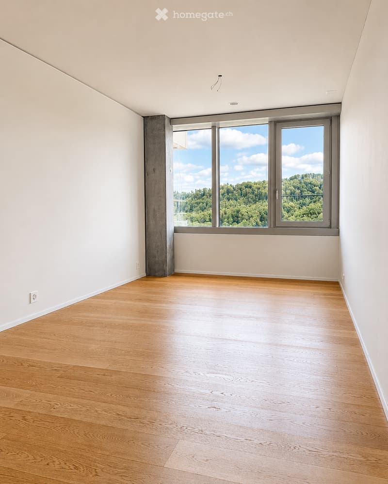 Empty room with light wooden flooring, white walls, and large windows offering a view of greenery outside.