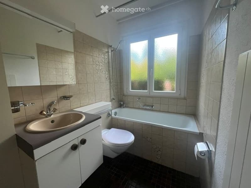 Bathroom with beige tiles, a white sink with a modern faucet, a toilet, a bathtub, and a shower. The room features a window with frosted glass and a white cabinet above the sink.
