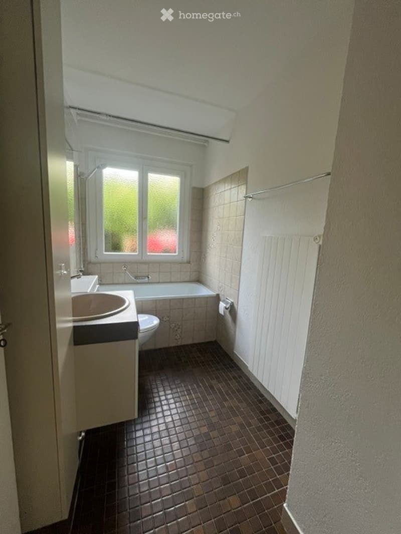 Bathroom with a dark tiled floor, a white sink, a bathtub, and a toilet. The room has a window with a view of greenery outside and a radiator on the wall.