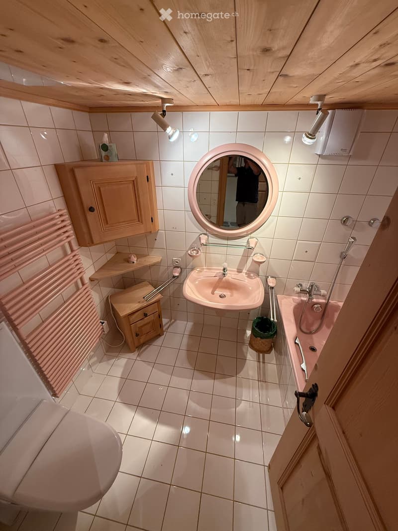 Bathroom with a pink sink, round mirror, pink bathtub, wooden cabinet, pink radiator, and a small wooden shelf.