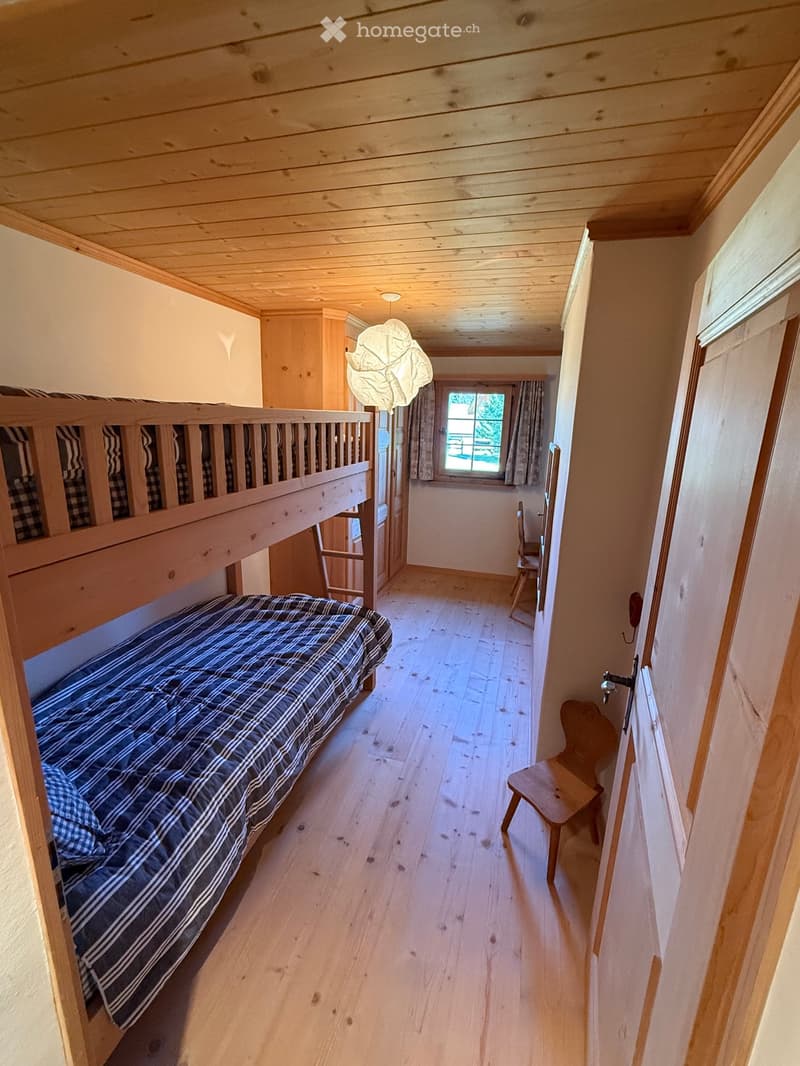 Cozy wooden bedroom with bunk bed, plaid bedding, wooden chair, and window with curtains.