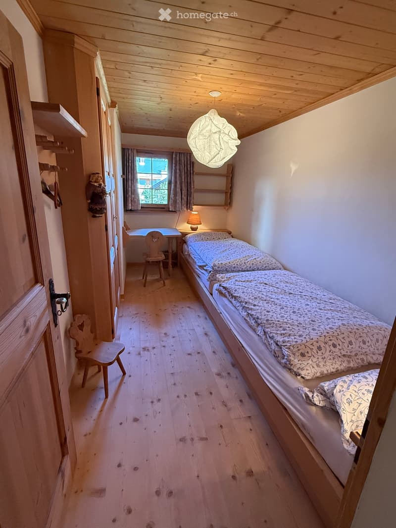 Cozy bedroom with wooden floors, two single beds, a small table with a chair, a lamp, and a window with curtains.