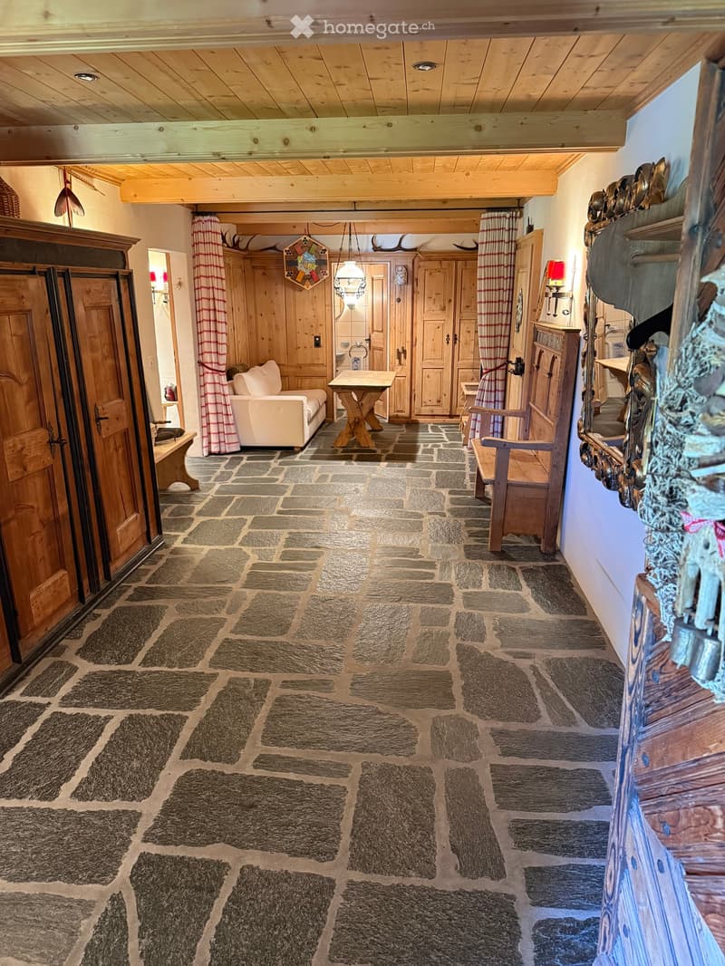 A rustic living room with wooden beams, stone flooring, a sofa, a small table, and a wooden chair. Wooden cabinets and a mirror are visible on the walls.