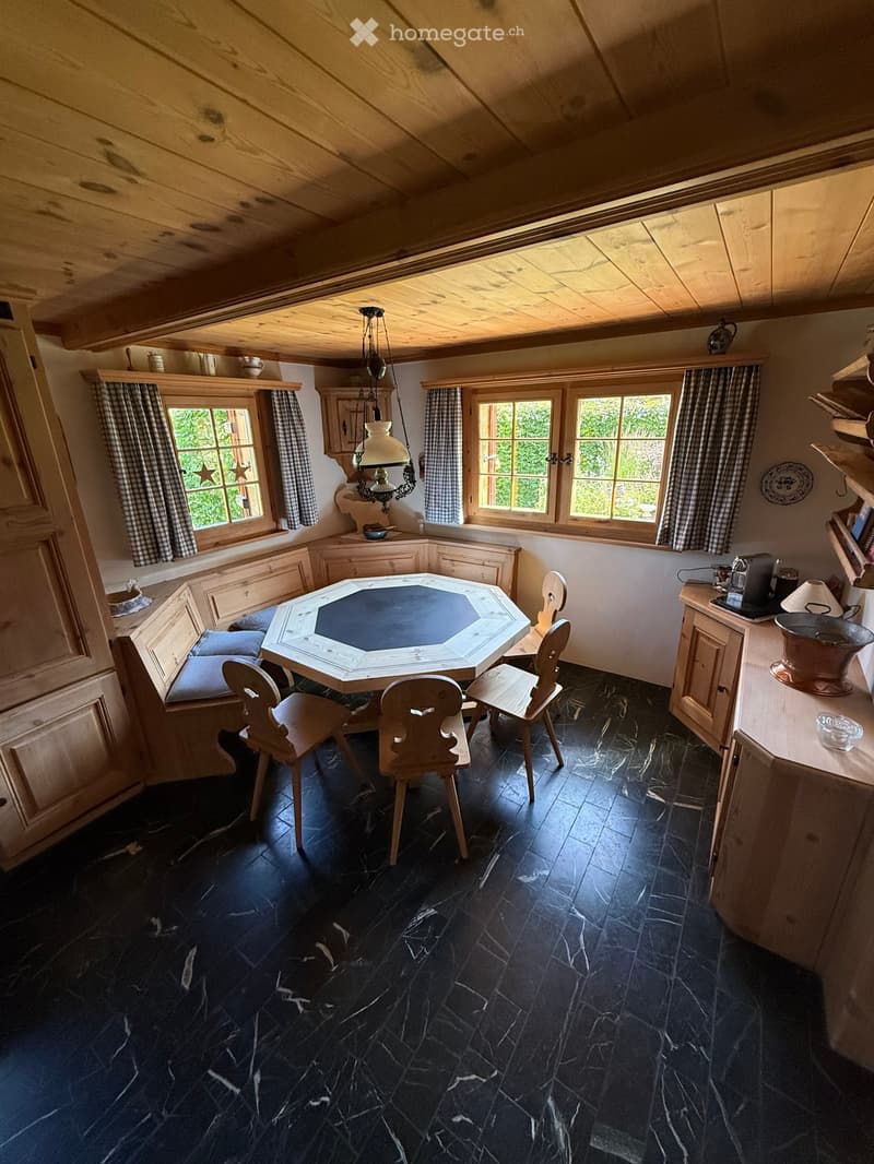 Cozy dining area with wooden beams, dark stone floor, octagonal table, wooden chairs, and built-in bench seating along the wall.