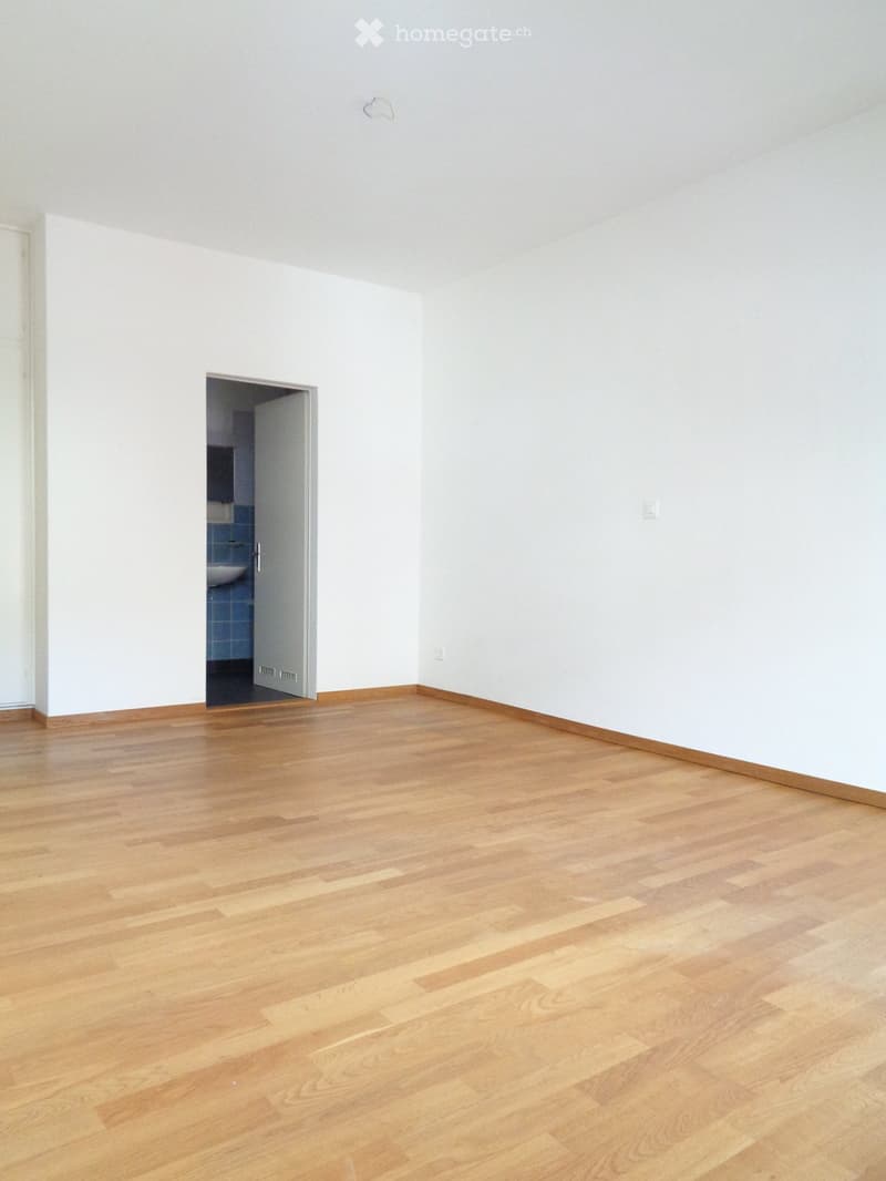 Empty room with light wooden flooring and white walls, featuring an open doorway leading to a small bathroom with blue tiles and a sink.