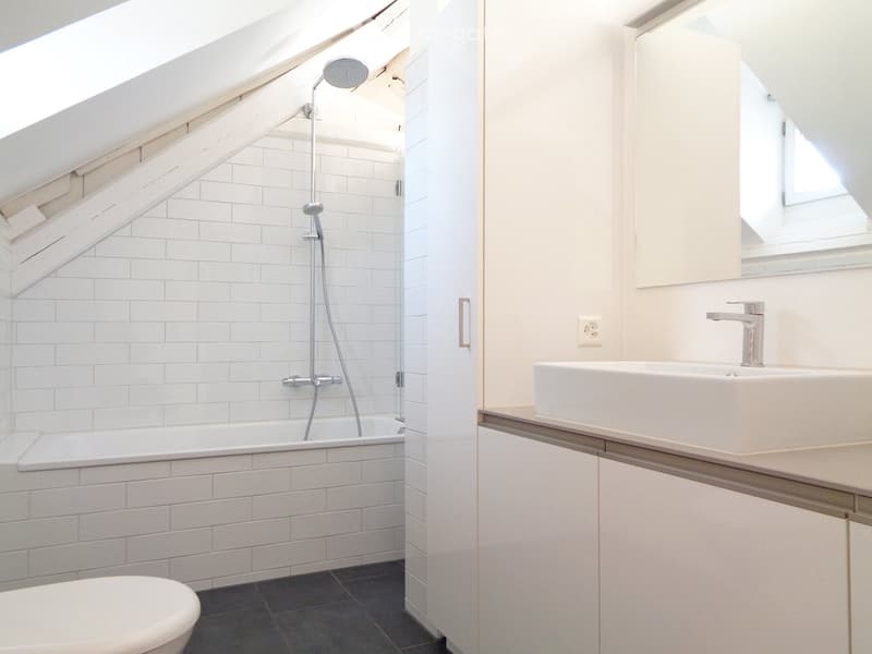 Modern bathroom with white tiled walls, a bathtub, toilet, sink, and a showerhead. Features a skylight window and sleek fixtures.