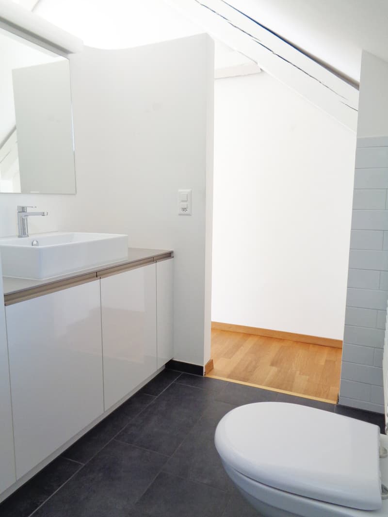 Modern bathroom with white sink, chrome faucet, mirror, white cabinet, and toilet. Dark tiled floor and light wooden flooring visible in adjacent area.