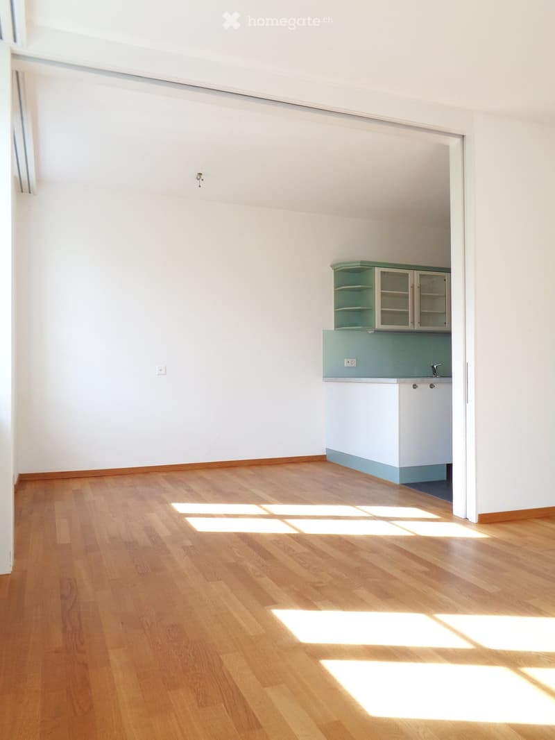 Empty room with light wooden flooring and white walls, featuring a small kitchen area with cabinets and a sink.