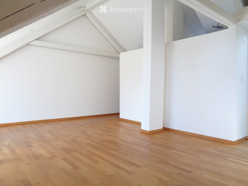 Empty room with white walls and wooden flooring, featuring exposed wooden beams on the ceiling and a small raised platform area.
