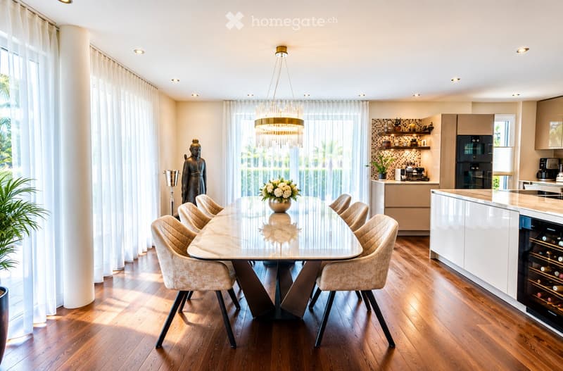 Open-plan dining and kitchen area with a modern glass dining table, six upholstered chairs, a chandelier, sleek kitchen cabinets, and a wine cooler. The space features large windows with sheer curtains and a statue near the window.