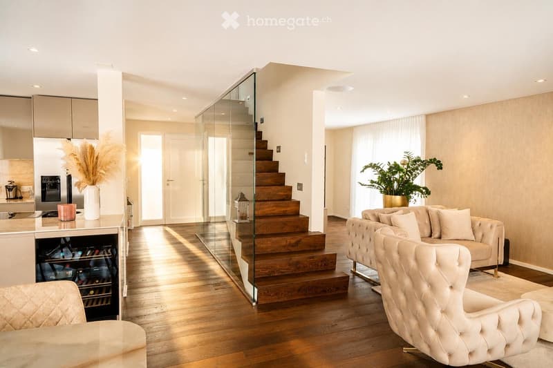 Modern living room with a cream-colored sofa, matching armchair, ottoman, wine cooler, coffee machine, decorative vase with dried flowers, candle, and a large potted plant. The space features wooden flooring, a glass staircase, and large windows allowing
