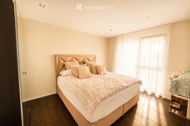 A well-lit bedroom with a large bed featuring a decorative headboard, multiple pillows, and a patterned blanket. The room has wooden flooring, light-colored walls, and a window with sheer curtains allowing natural light to fill the space. A cat tree is po