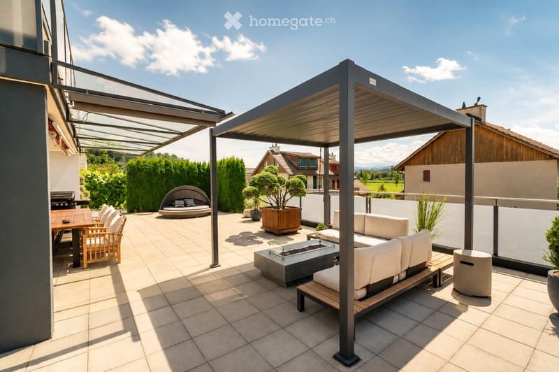 A modern outdoor patio with a covered seating area, featuring a sofa, wooden table, chairs, a fire pit, and potted plants.