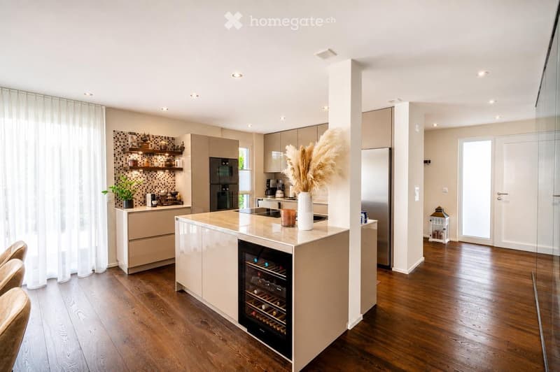 Modern kitchen with light wood flooring, white cabinets, a central island with a wine cooler, and built-in appliances including a refrigerator and oven. Shelves hold decorative items and a potted plant, with a vase and candle on the island.