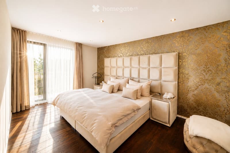 Elegant bedroom with a large upholstered headboard, a neatly made bed with multiple pillows, a nightstand with a lamp, and a small stool near the window.