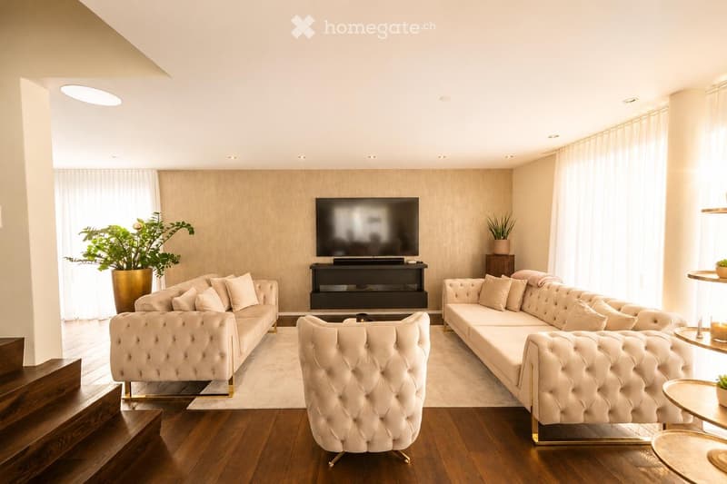 Modern living room with light-colored walls, wooden flooring, and large windows. Features a cream-colored sofa, matching armchair, coffee table, television on a stand, and decorative plants.