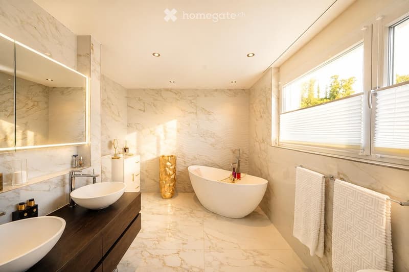 Modern bathroom with marble walls and floors, a large bathtub, double sink vanity, and a large mirror. The space features recessed lighting and a window with blinds.