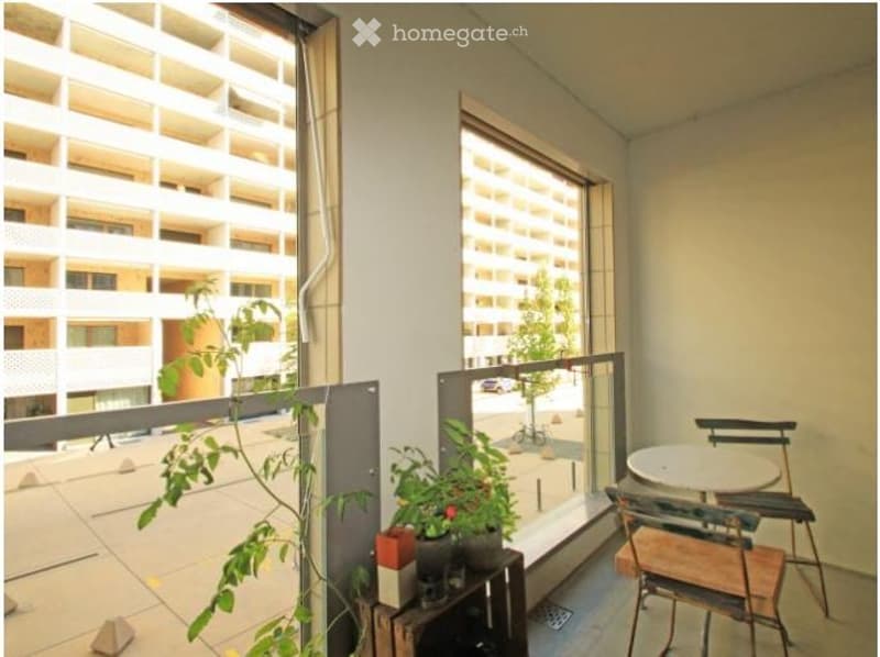 Balcony with a view of a modern apartment building, featuring a small table, chairs, and potted plants.