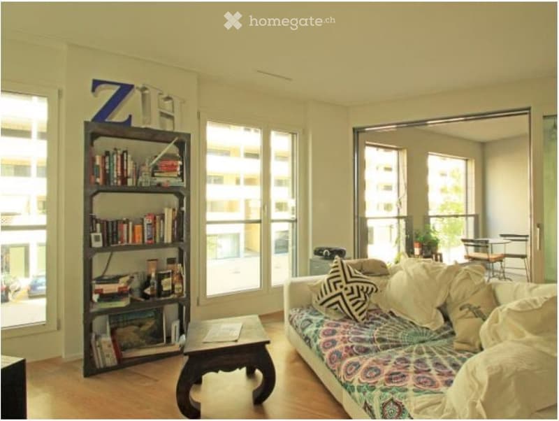 Bright living room with large windows offering a city view, featuring a colorful patterned sofa, wooden coffee table, bookshelf filled with books, and a small dining area with a table and chairs.