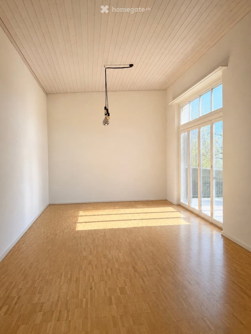 Empty room with wooden flooring, white walls, and large windows allowing natural light to enter. A single pendant light fixture hangs from the ceiling.
