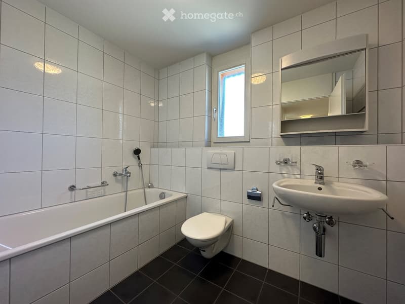 Modern bathroom with white tiled walls and dark tiled floor, featuring a bathtub, toilet, sink, mirror, and showerhead.