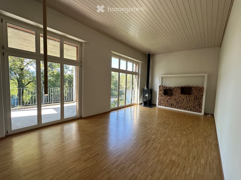 Bright, unfurnished room with wooden flooring, large windows, and a fireplace area with stacked firewood.
