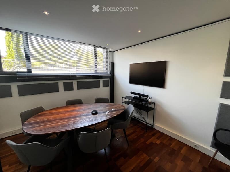Modern conference room with a round wooden table, several chairs, a large screen, and a small shelf with electronic devices.